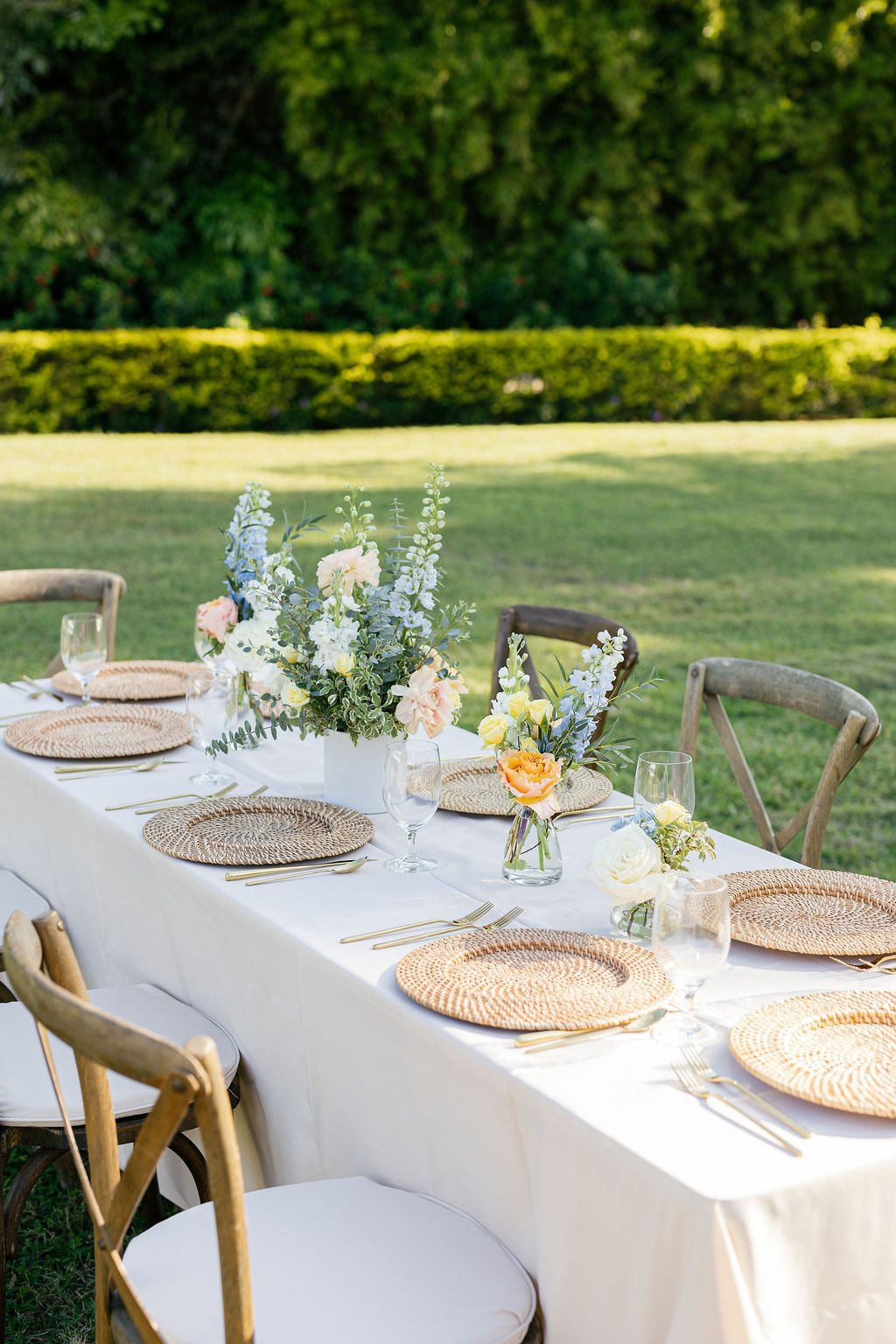 An outdoor dining table set with woven placemats, glassware, and floral centerpieces in a lush green garden.