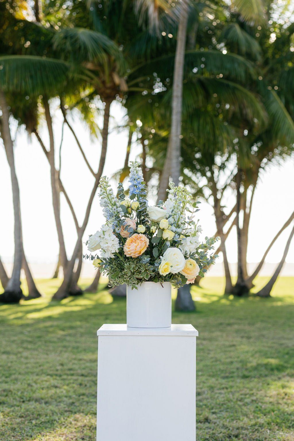 A large bouquet of assorted flowers, including roses and other blooms, arranged in a white vase on a white pedestal outdoors with palm trees in the background.