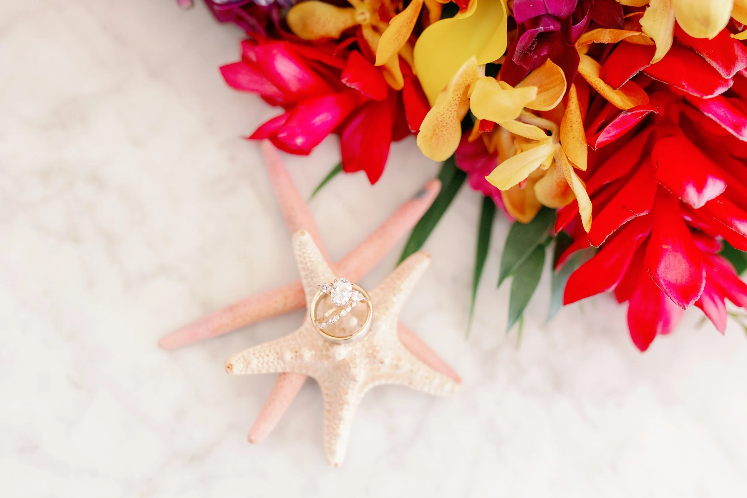 Starfish with an engagement ring, surrounded by vibrant red, yellow, and purple flowers on a marble surface.