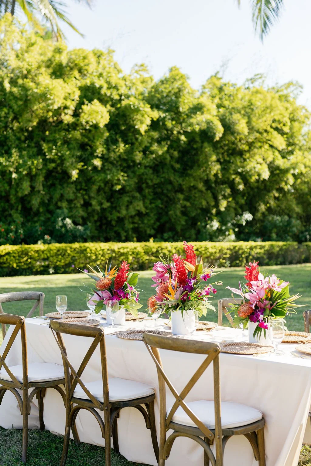 Outdoor table set for a meal with floral centerpieces, glasses, and woven placemats on a white tablecloth, surrounded by wooden chairs, in a lush garden setting with green trees and shrubbery in the background.