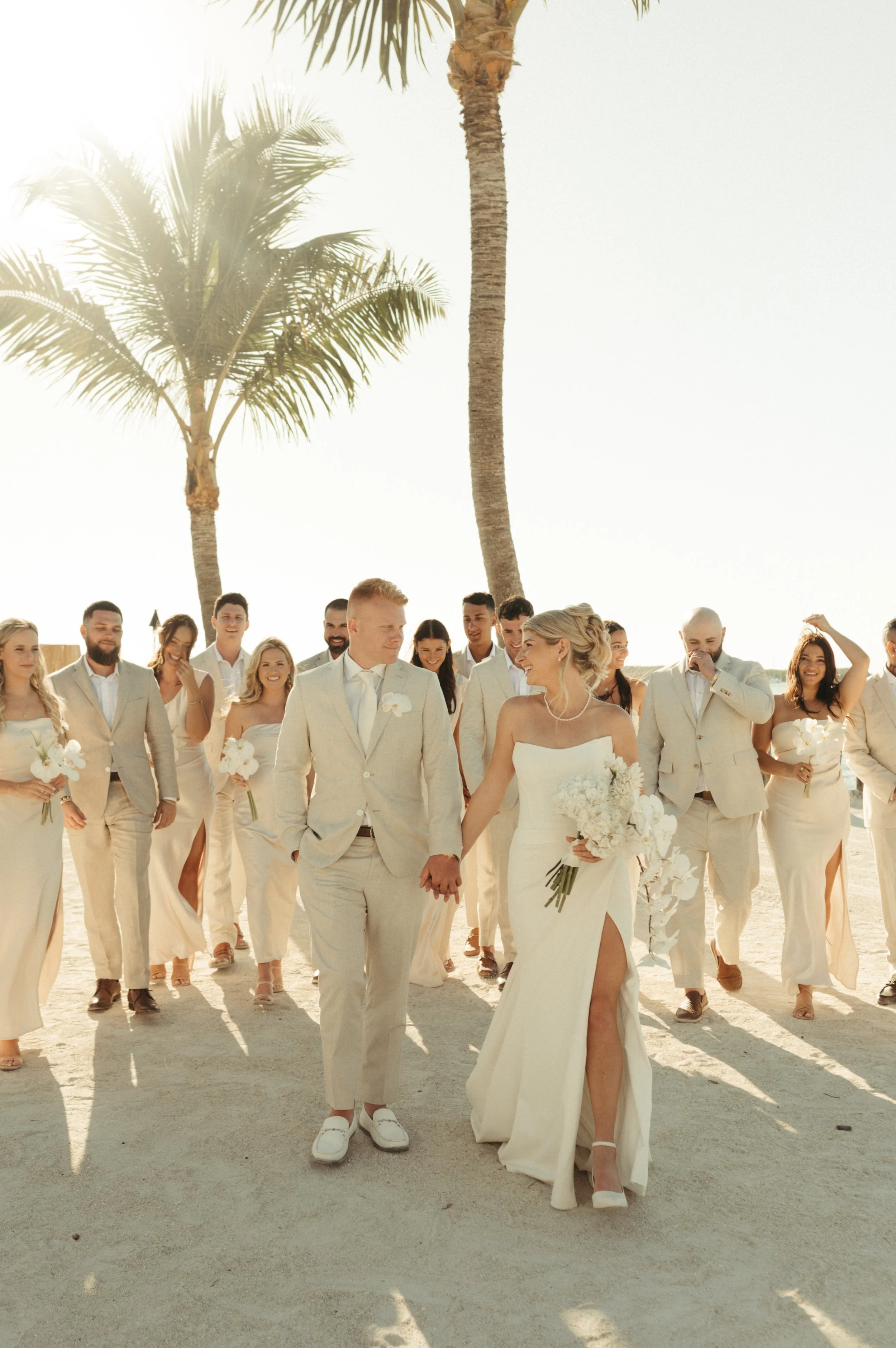 Beach wedding party in light attire walking with palm trees in background.