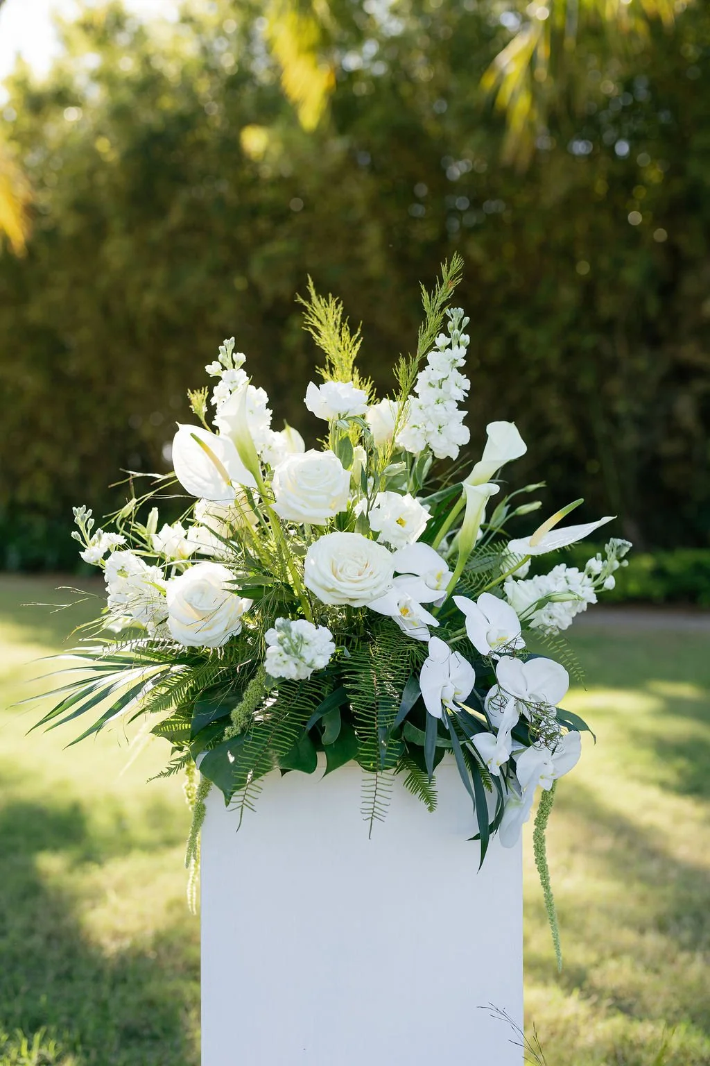 White floral arrangement with roses and calla lilies, placed outdoors on a white stand with greenery in the background.