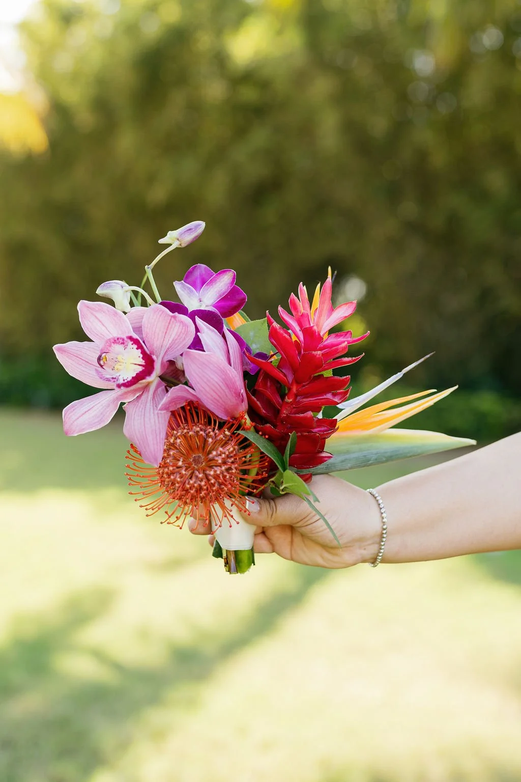 A person's hand holding a vibrant bouquet of various colorful flowers against a blurred green outdoor background.