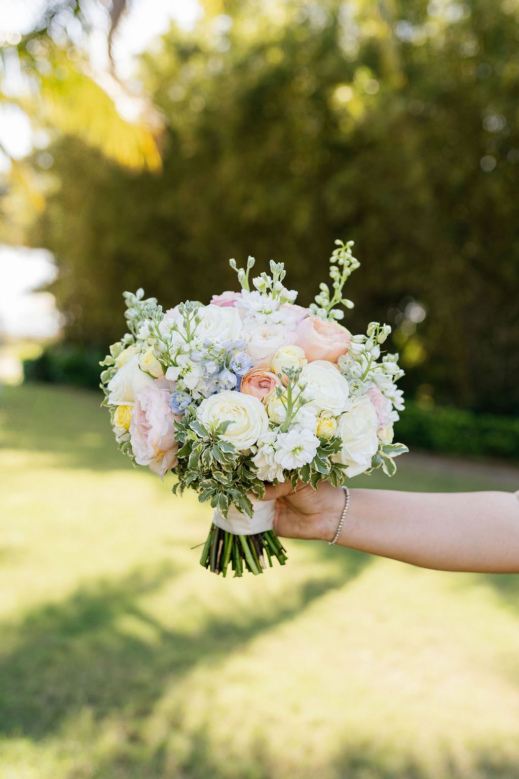A person holding a large bouquet of white, pink, and peach flowers with greenery outdoors on a sunny day.