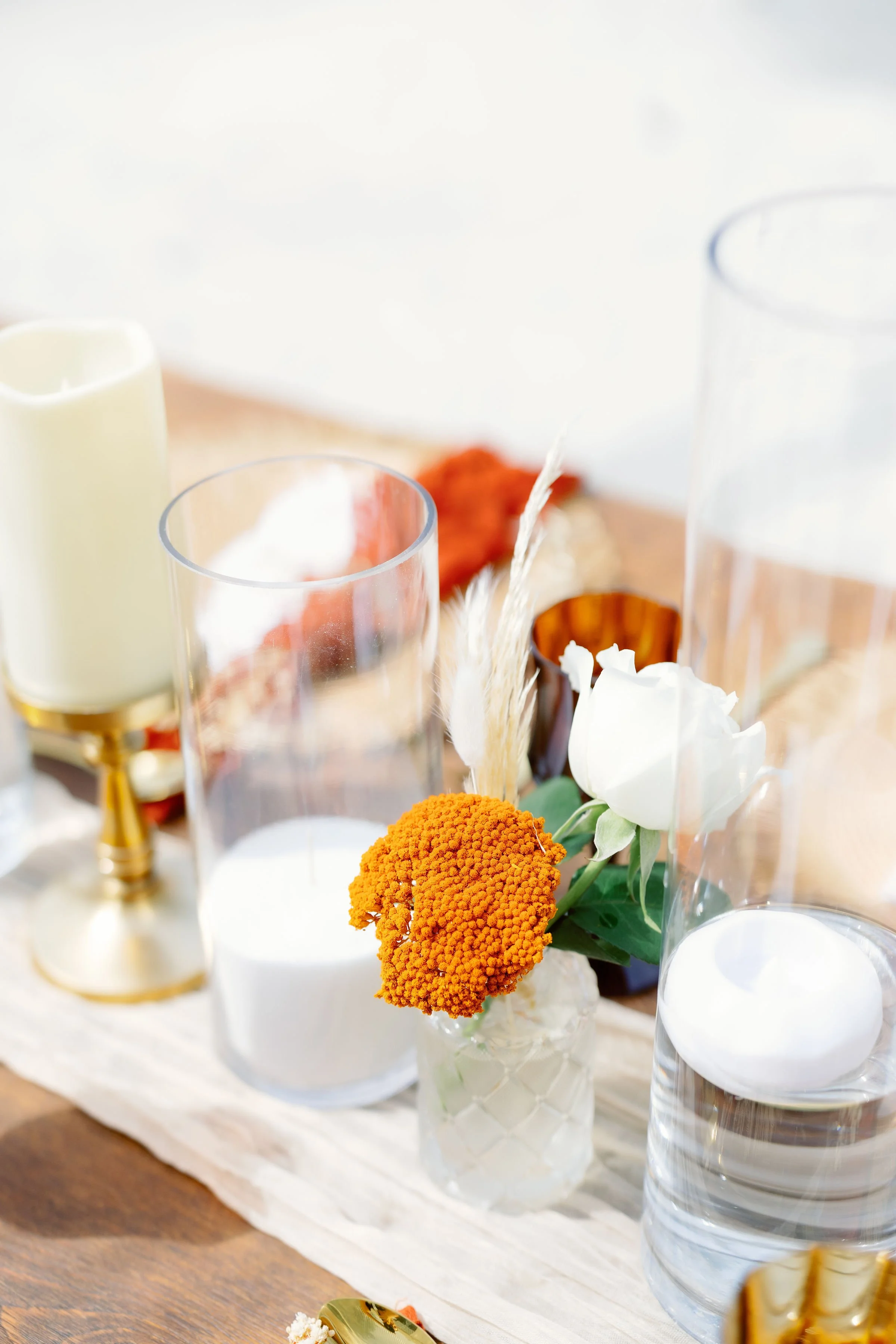 Decorative table setting with candles, glass holders, orange and white flowers in a vase, and a golden candlestick on a wooden table.
