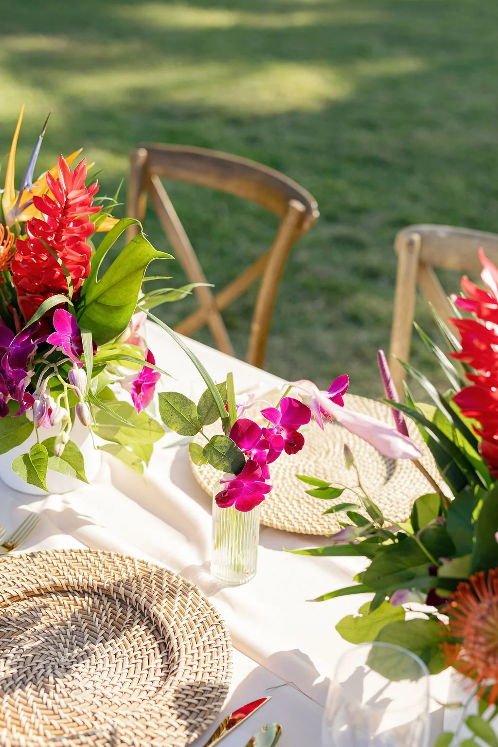A table with a white tablecloth decorated with vibrant pink and red tropical flowers and green leaves, set outdoors on a sunny day with grass and trees in the background.