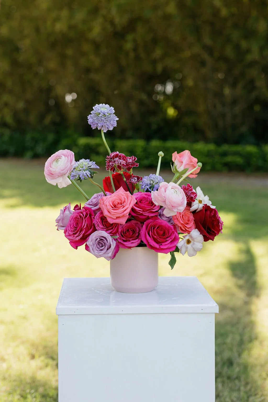 Vase with pink, purple, red, white, and lavender flowers on a white pedestal outdoors with a blurred green background.
