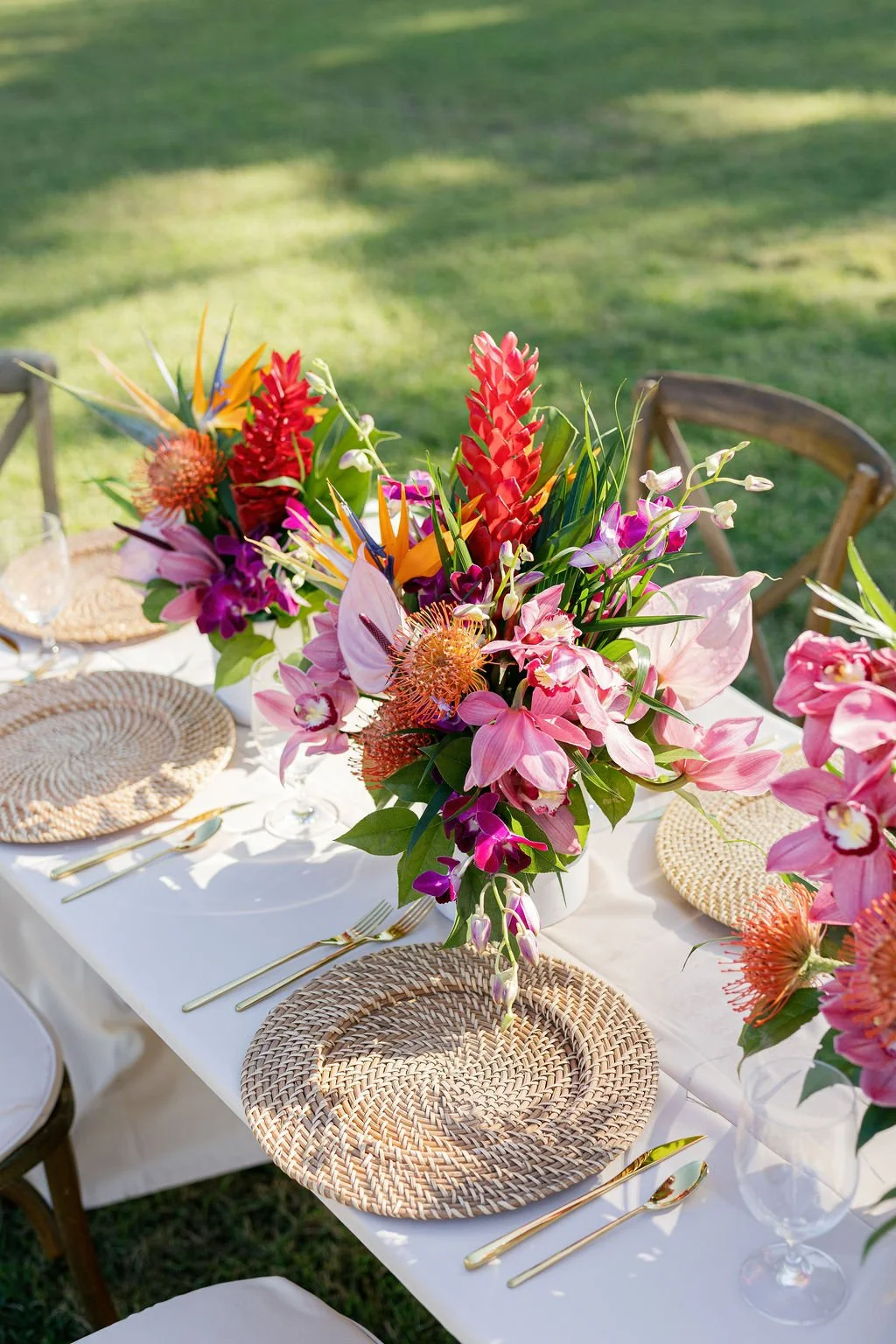 Colorful floral centerpiece on a table set outdoors with woven placemats, gold cutlery, and clear wine glasses.