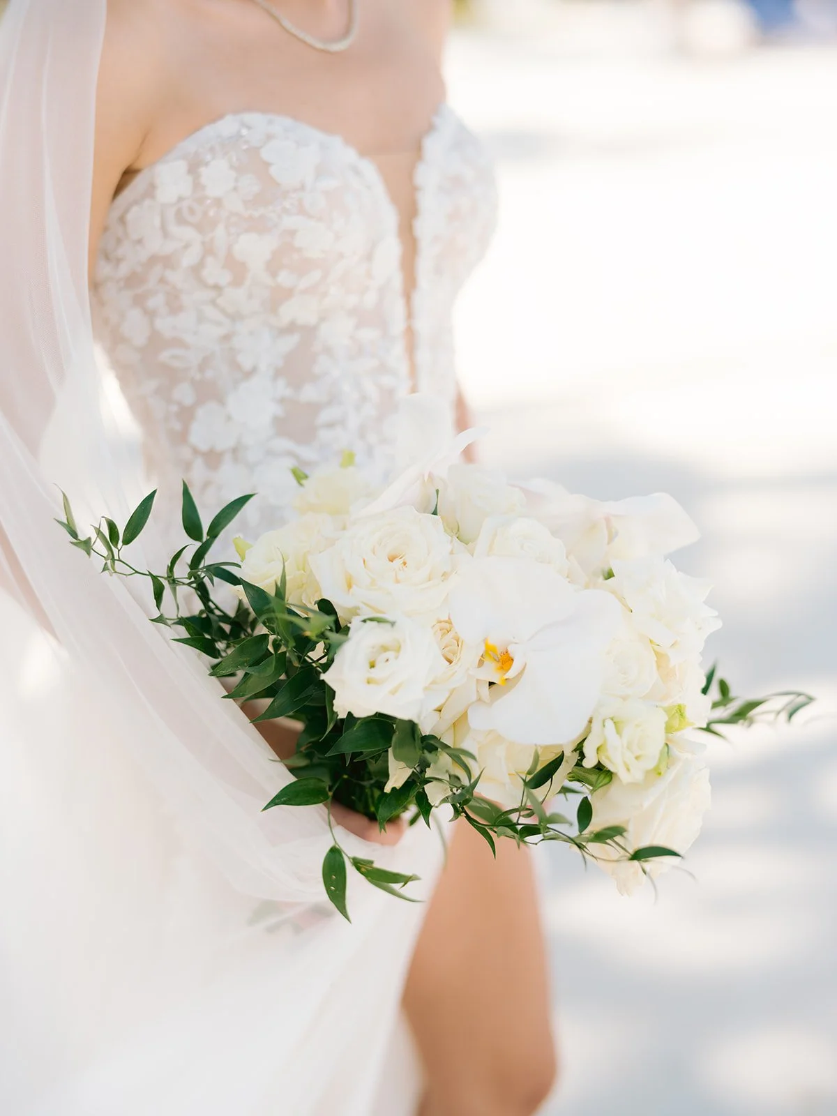 Bride holding a bouquet of white roses and orchids, wearing a lace wedding dress with floral patterns.