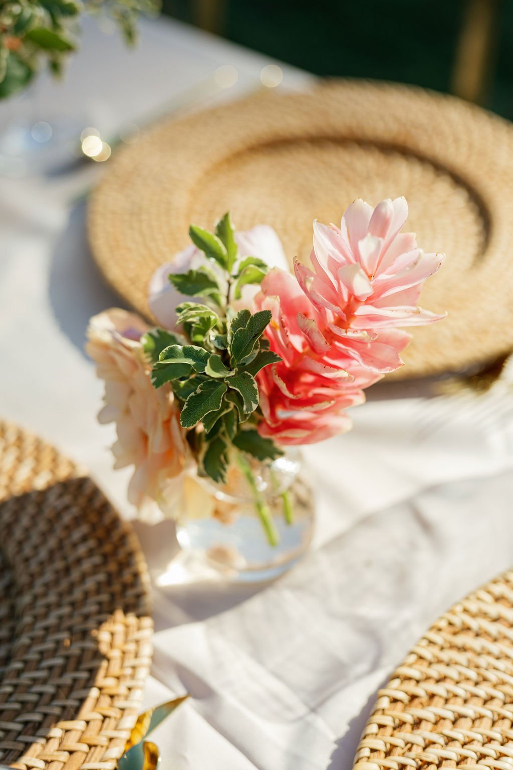 Pink and white flowers in a glass vase on a table with a woven straw hat and wicker placemats.