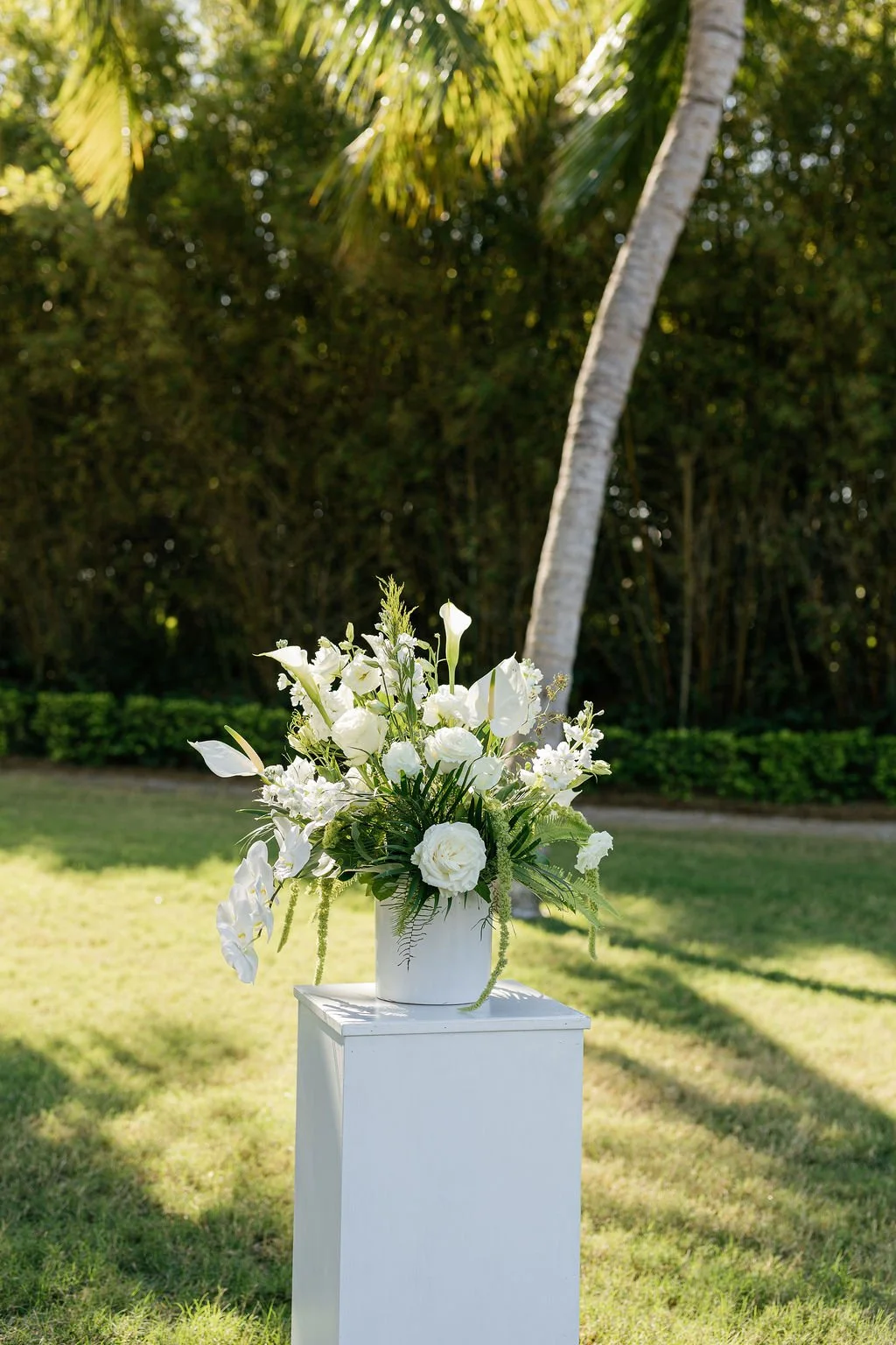 White floral arrangement in a white vase on a white pedestal outdoors with greenery and trees in the background.