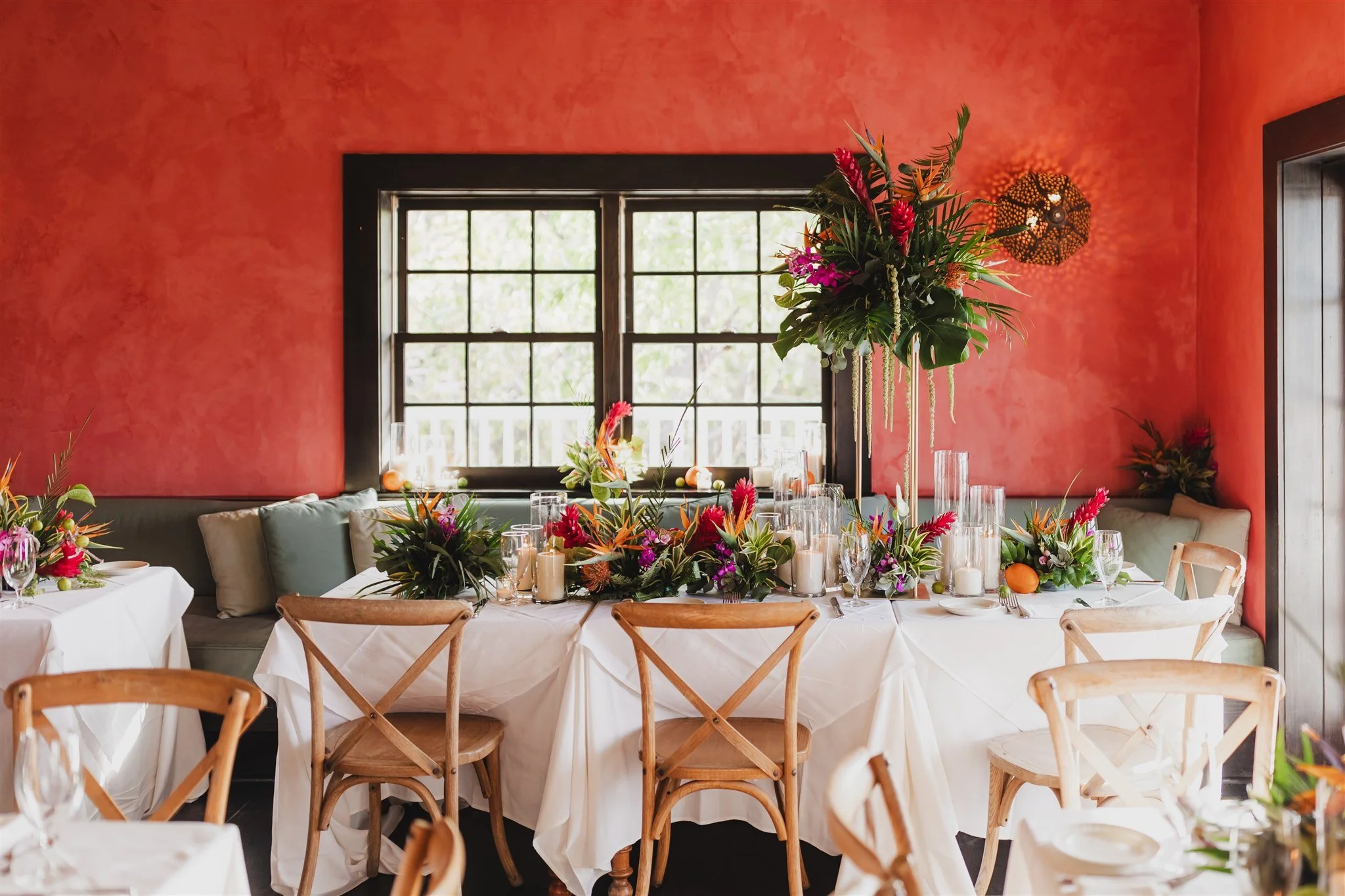 Dining room with vibrant red walls, tables decorated with white tablecloths, tropical floral centerpieces, candles, and wooden chairs.