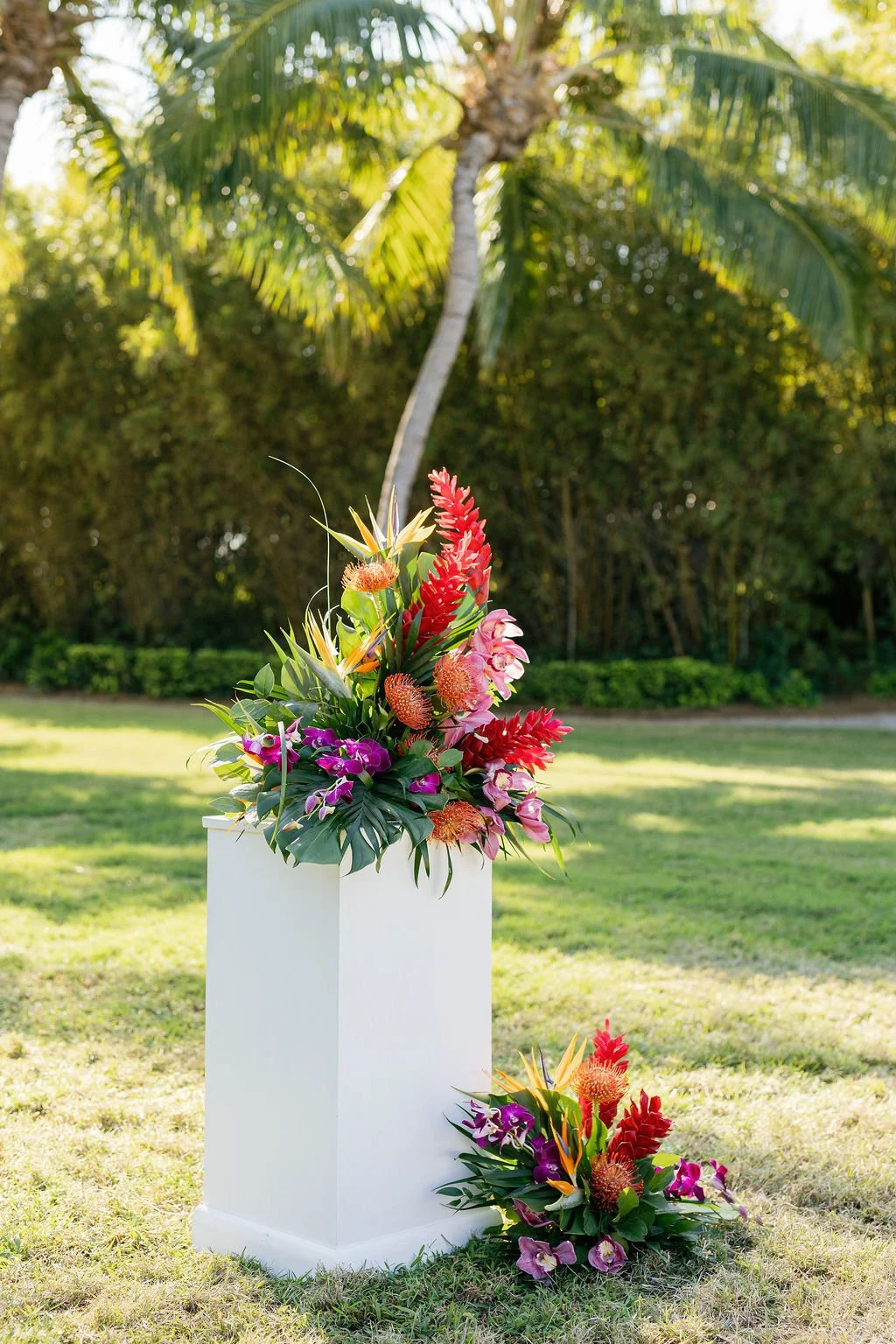 Colorful tropical flower arrangement on a white pedestal outdoors with palm trees in the background.