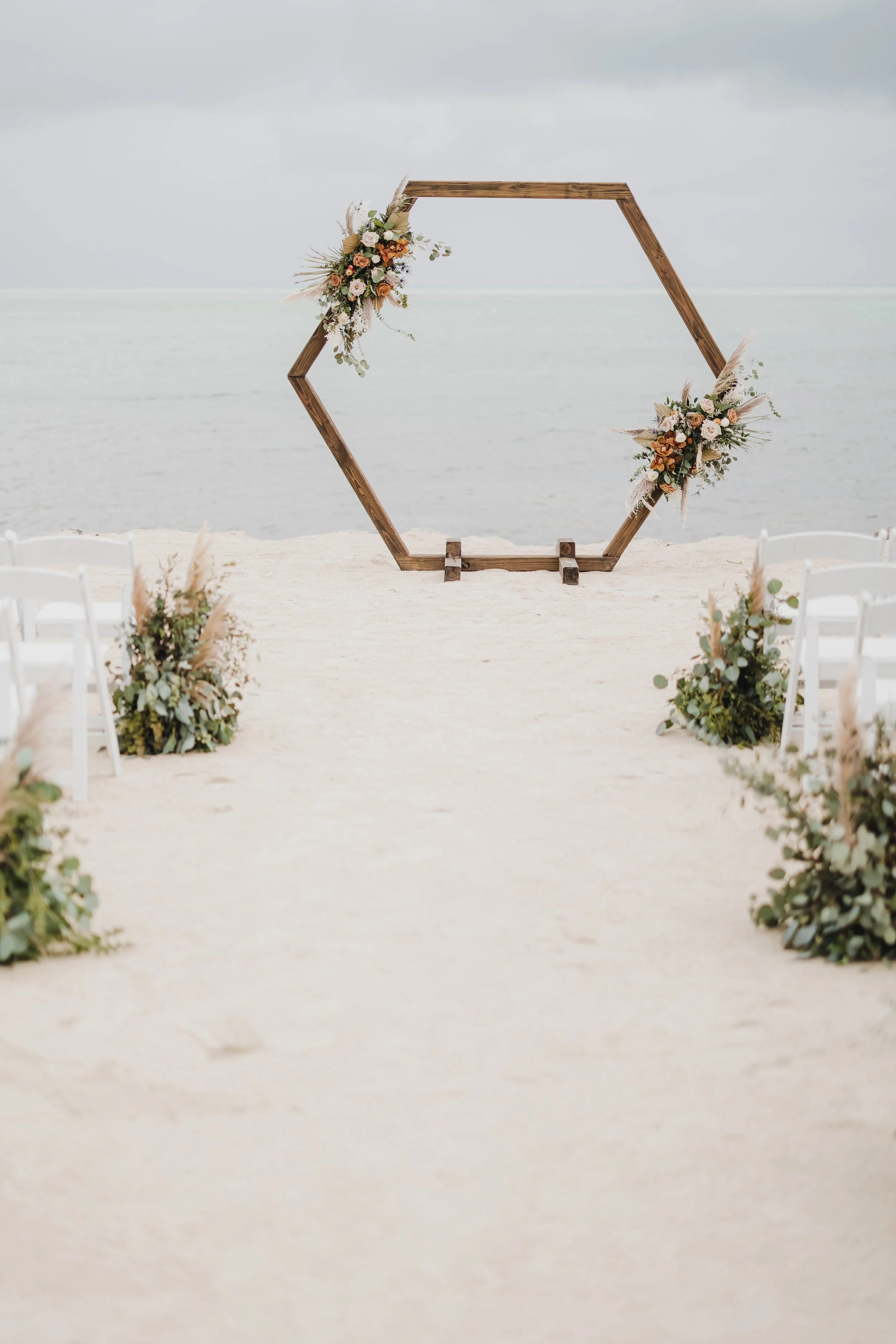 Beach wedding setup with hexagonal wooden arch decorated with flowers, surrounded by white chairs and greenery, overlooking the ocean.