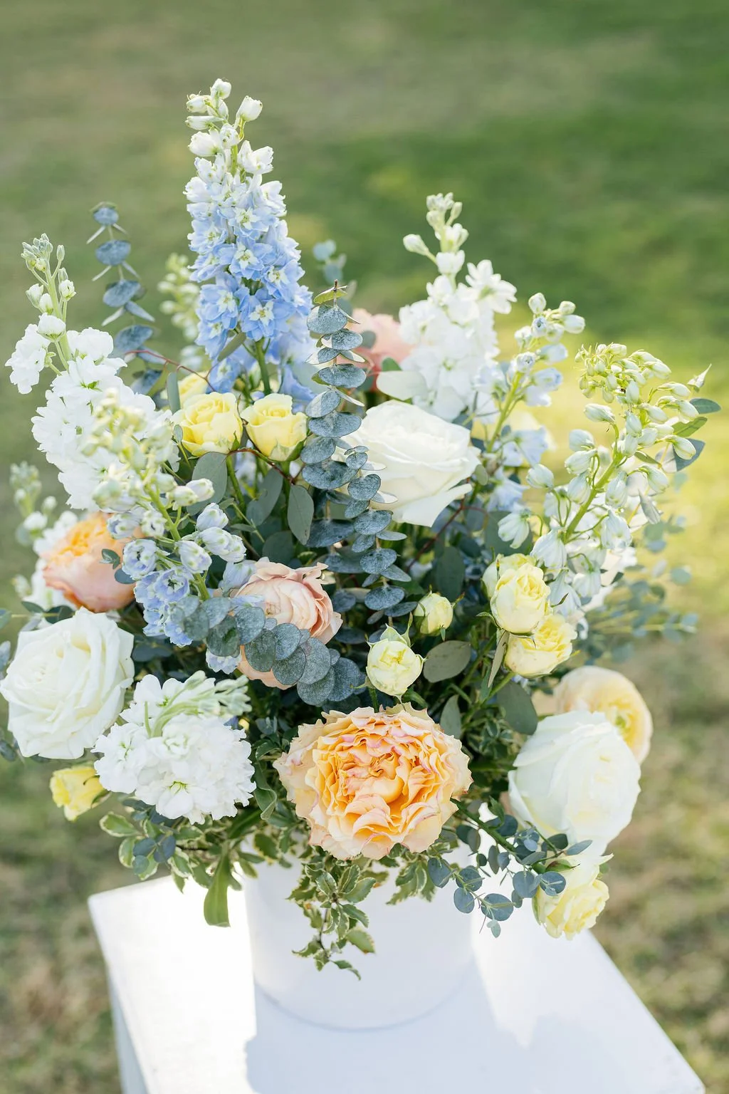 A floral arrangement with white, yellow, and peach roses, blue delphiniums, green eucalyptus, and other greenery in a white vase outdoors on a sunny day.