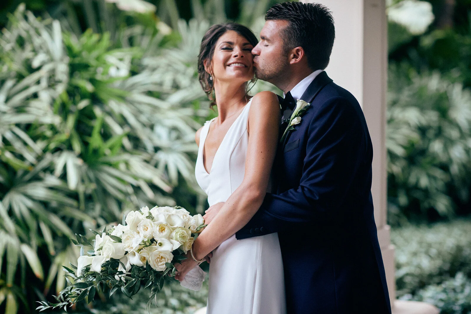 A bride in a white dress holding a bouquet of white flowers while a groom in a navy suit kisses her on the cheek during a wedding ceremony, surrounded by lush green foliage.