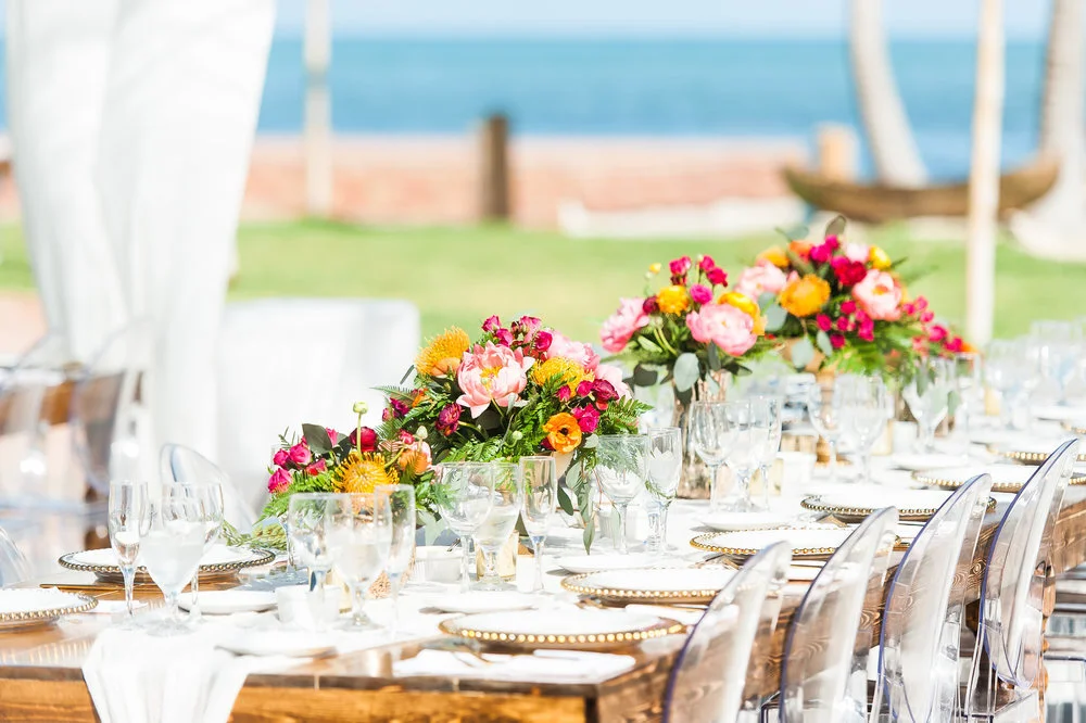 Outdoor dining table with floral centerpieces and glassware near the water