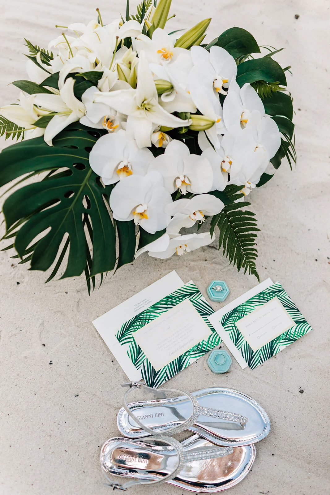 White floral bouquet with lilies and orchids on sand, alongside wedding invitations with tropical leaf prints, a jewelry ring in a blue box, and silver sandals.