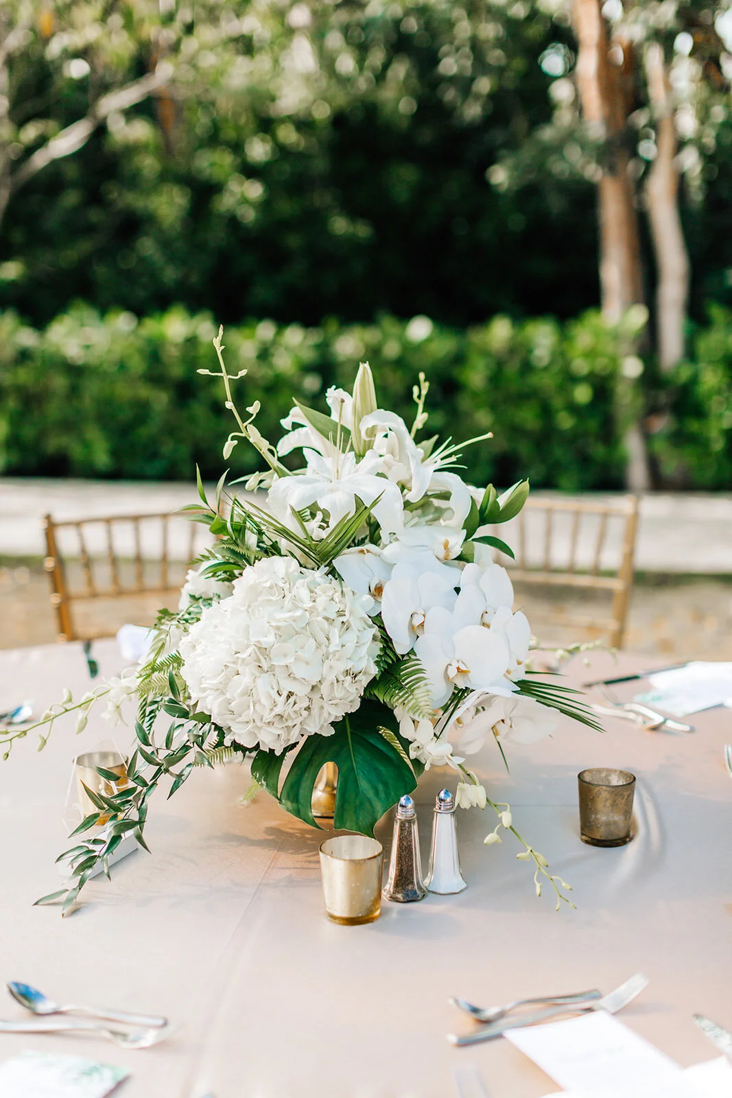 Elegant floral centerpiece with white lilies, orchids, and hydrangeas on an outdoor dining table, surrounded by gold votive candles and silver salt and pepper shakers.