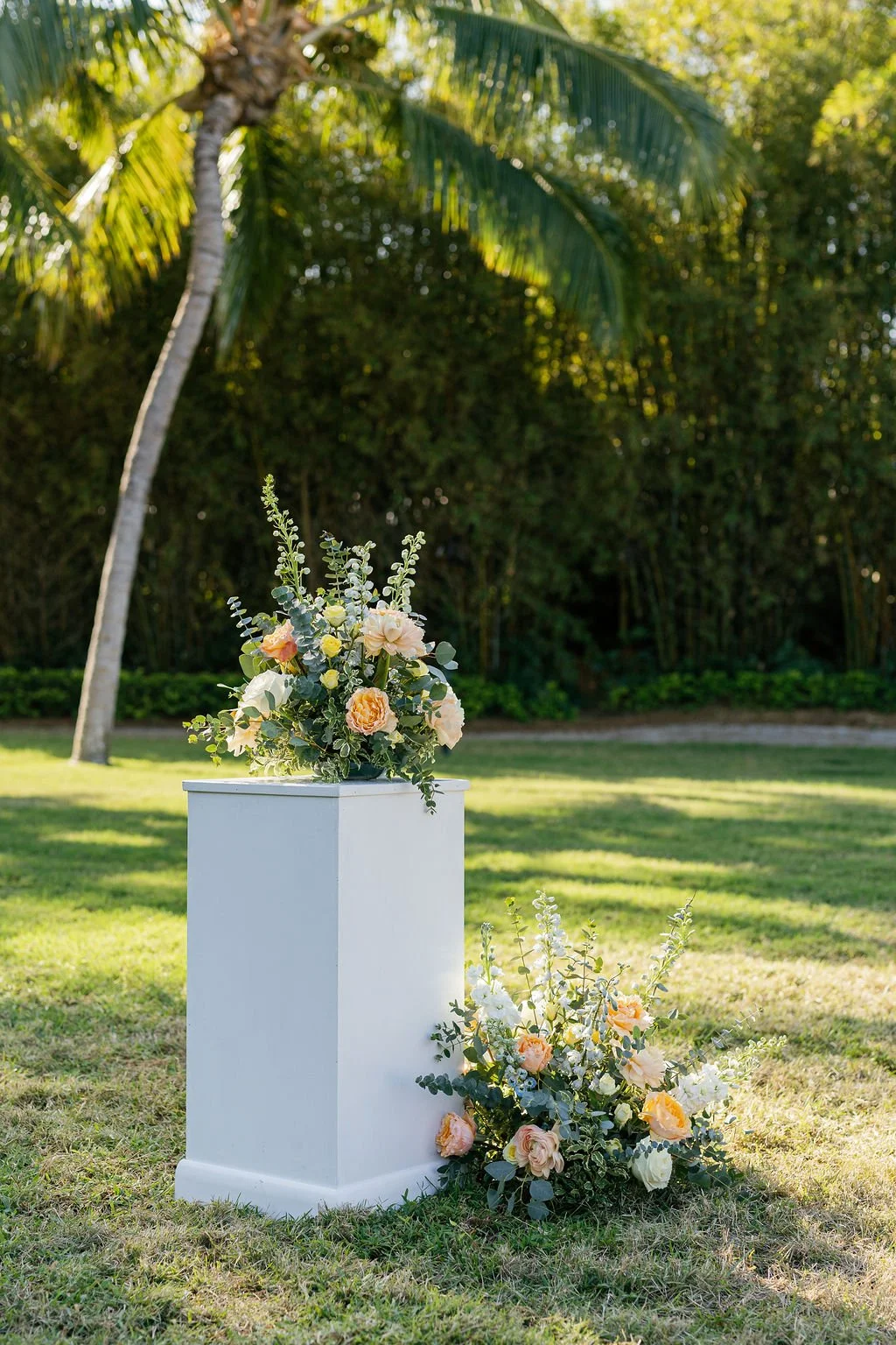 A floral arrangement on a white pedestal outdoors with green trees and grass in the background.