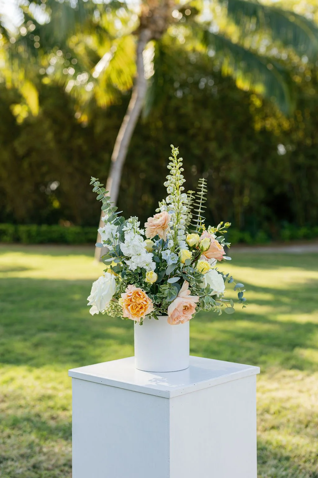 A white flower vase with pastel-colored roses and white flowers, placed on a white pedestal outdoors with green grass and trees in the background.