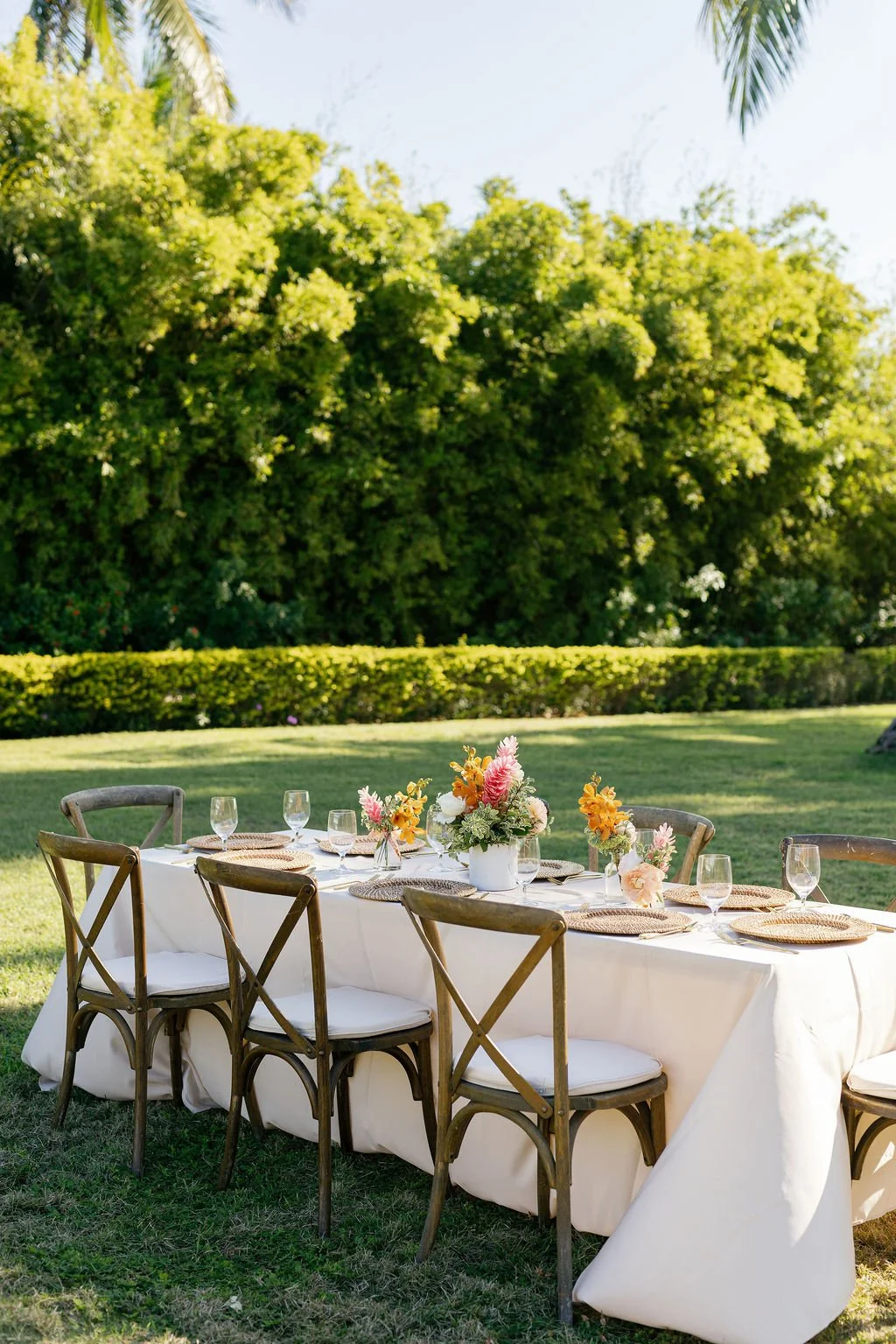 An outdoor dining table set for a meal with floral centerpieces, surrounded by wooden chairs, on a grassy lawn with green trees in the background.