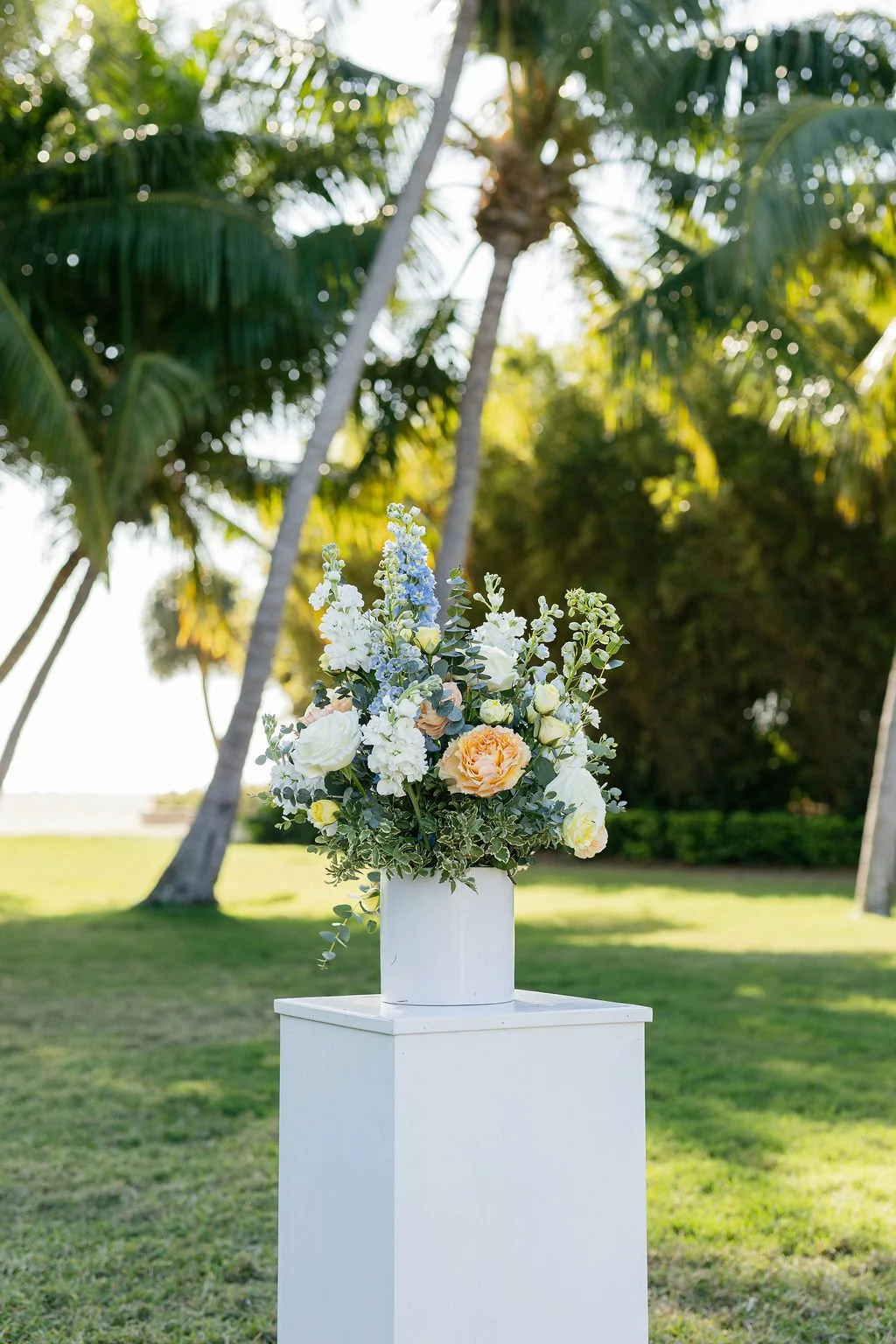 A floral arrangement in a white vase on a white stand outdoors with palm trees and green grass in background.