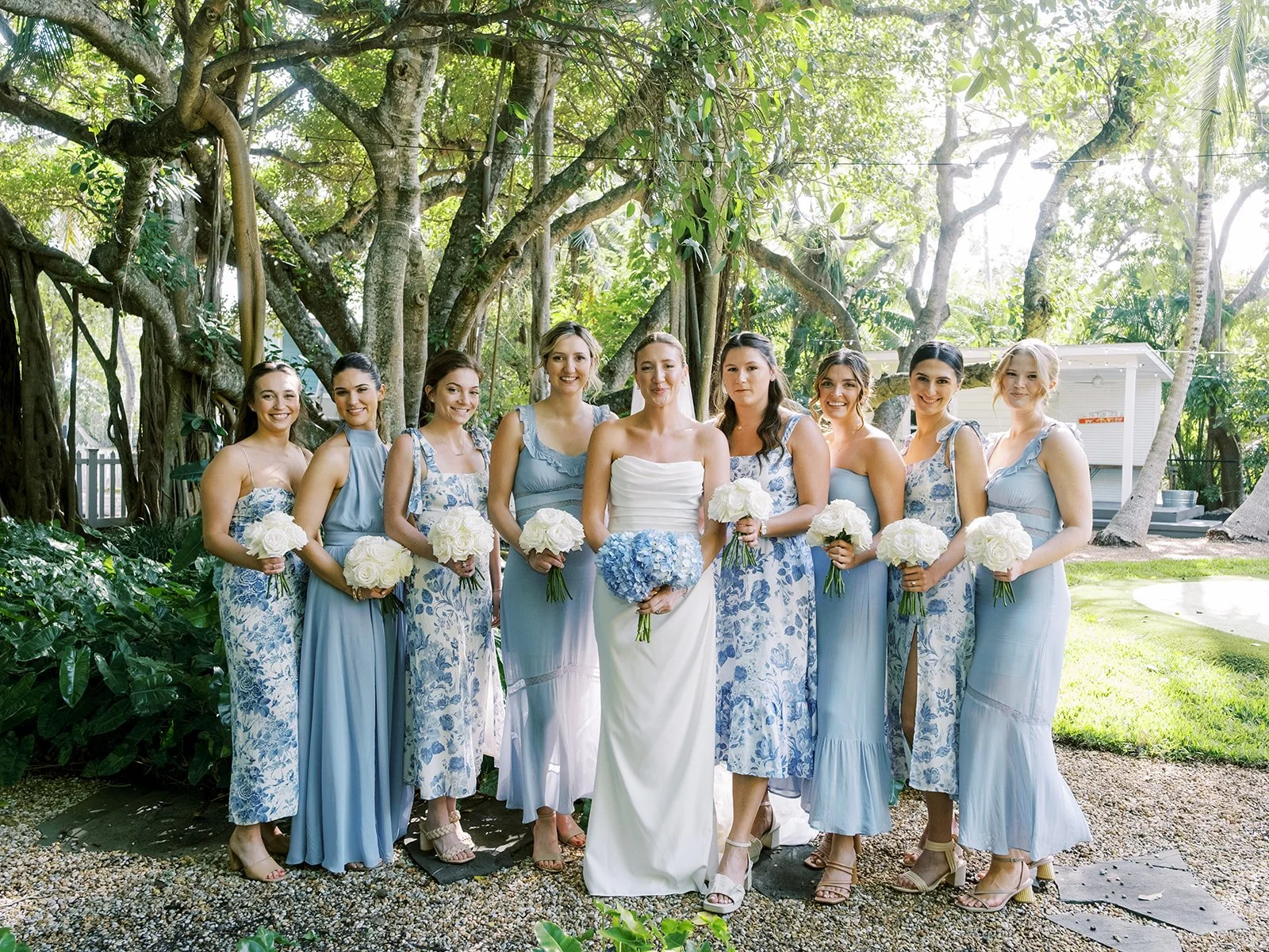 A bride in a white dress holding a bouquet stands with eight bridesmaids in blue and white dresses, each holding white bouquets. They are outdoors in a lush garden setting with trees and greenery.