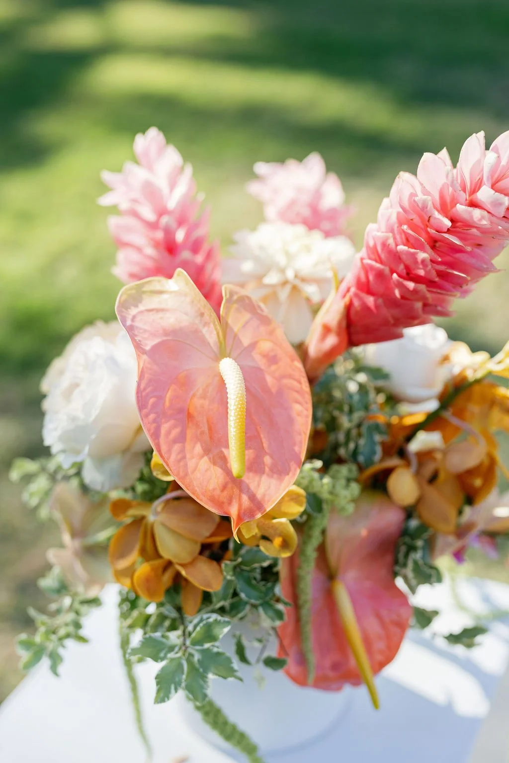 A vibrant bouquet of pink, white, and orange flowers with lush green foliage, featuring an anthurium with a yellow spadix and pink spathe, set against a blurred outdoor green background.