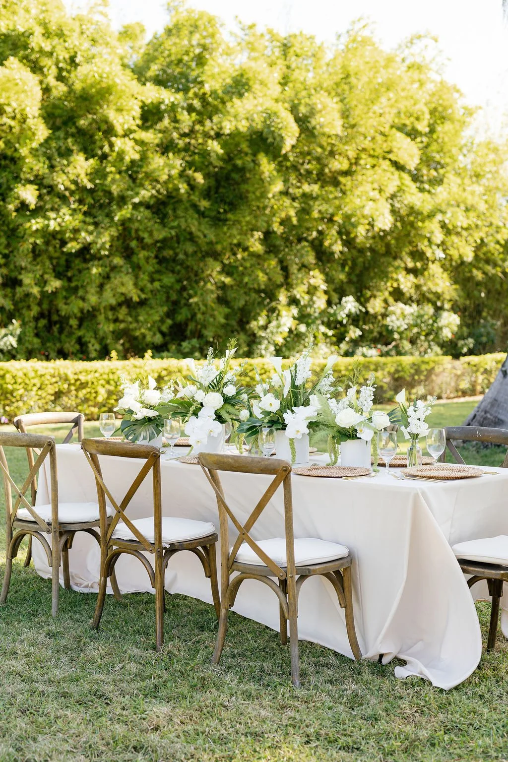 Outdoor wedding reception table decorated with white flowers, glasses, and plates, set amidst lush green trees and hedges