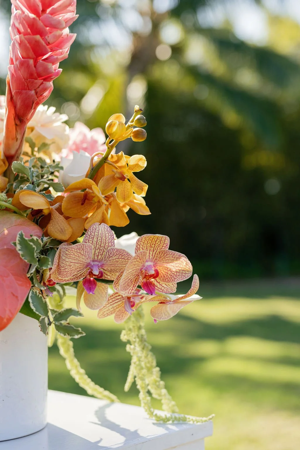 Close-up of a colorful flower arrangement with pink, yellow, and white flowers, including orchids, in a white vase outdoors.