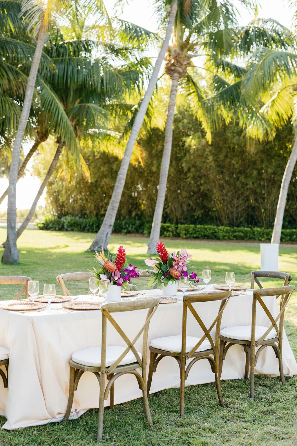 Outdoor table set for a gathering with napkins, glassware, and floral centerpieces, surrounded by wooden chairs, beneath tall palm trees on a sunny day.