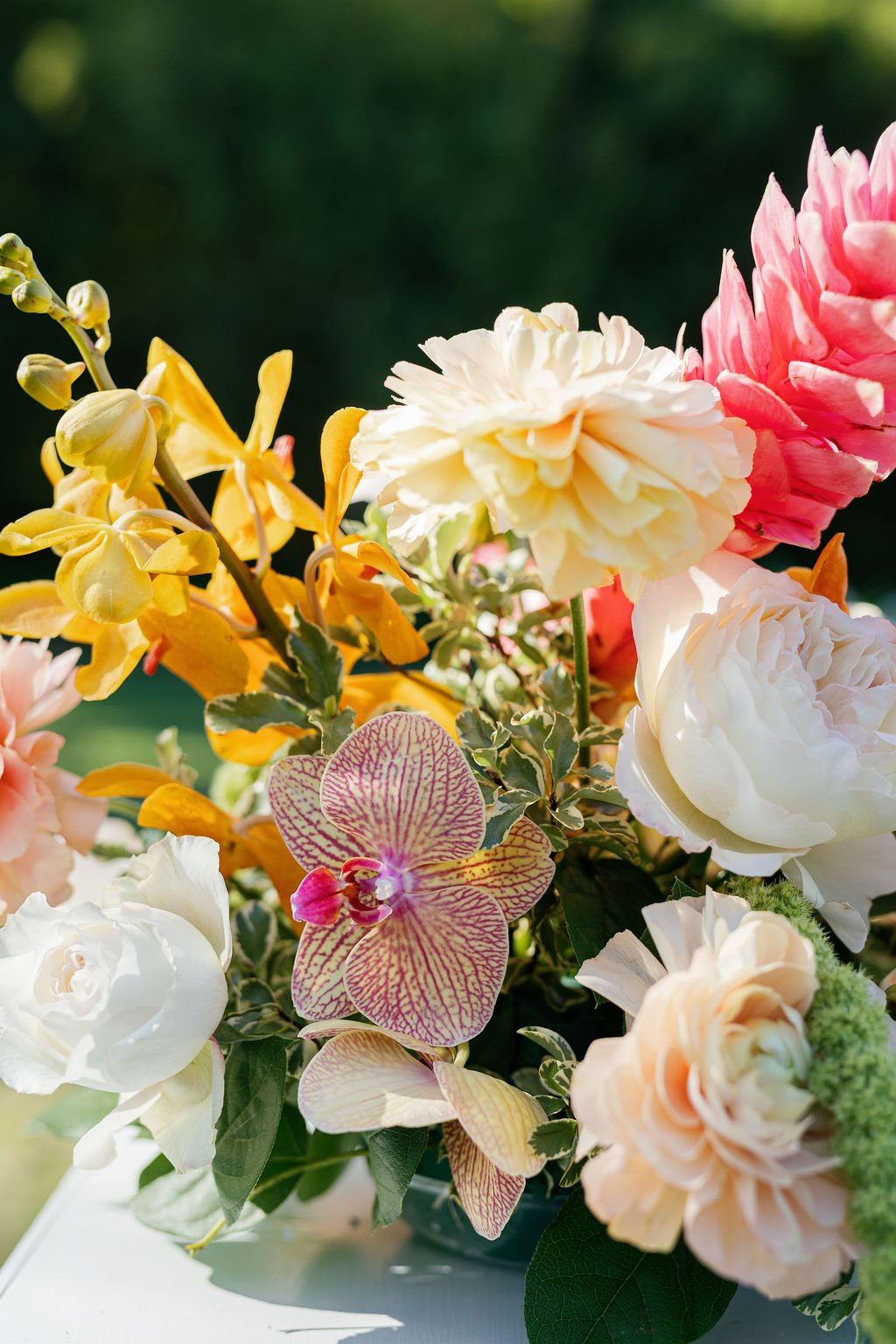 A vibrant bouquet of various flowers, including pink, white, yellow, and spotted orchids, with green leaves, set against a blurred dark green background.