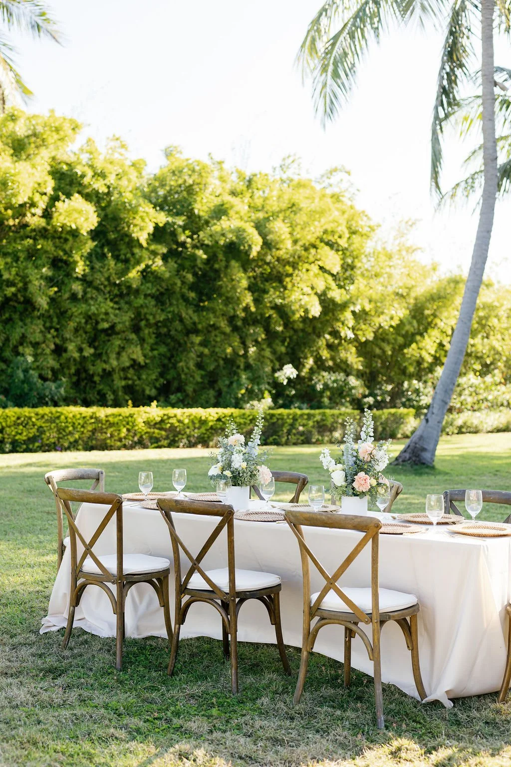 An outdoor banquet table set for a gathering, decorated with floral centerpieces, wine glasses, and placemats, surrounded by wooden chairs on a lush green lawn with trees in the background.