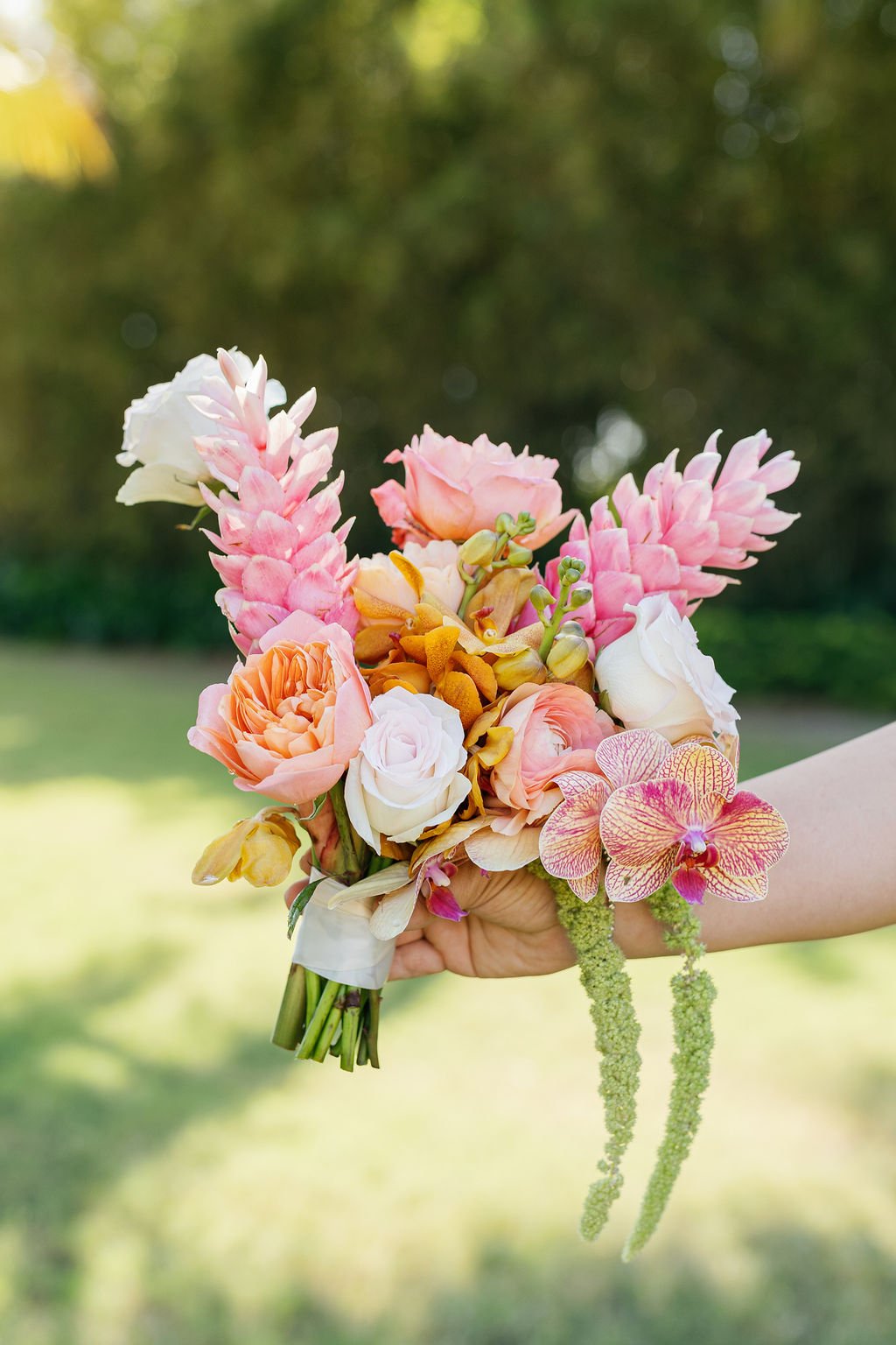 Close-up of a colorful flower bouquet held by a person against a blurred outdoor background. The bouquet includes pink, white, and peach roses, pink and white orchids, and other exotic flowers.