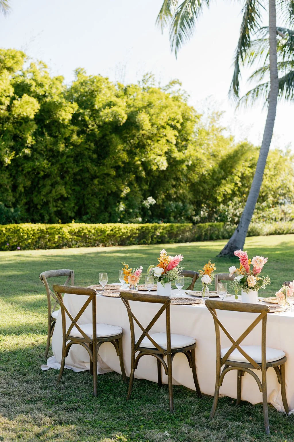 Outdoor garden party setup with a long table decorated with floral centerpieces, glasses, and tableware, surrounded by wooden chairs, lush green trees, and grass on a sunny day.