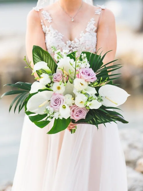 A bride in a lace wedding dress holding a large bouquet of white calla lilies, pink roses, and various green leaves and foliage.