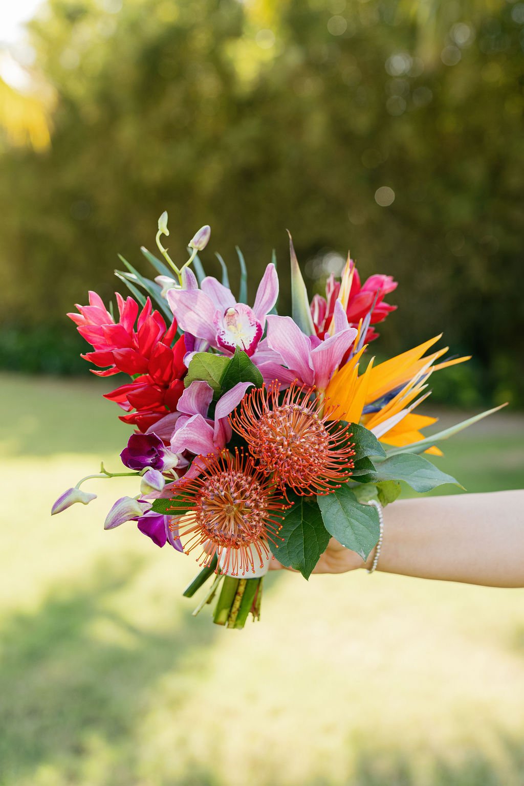A colorful bouquet of various flowers, including pink orchids, red ginger, orange protea, and purple flowers, held by a person's hand outdoors with a blurred green background.