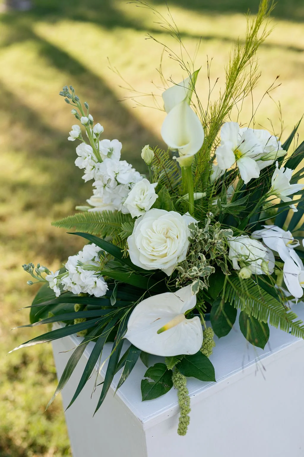 A floral arrangement with white calla lilies, roses, and various greenery, set against a blurred outdoor background.