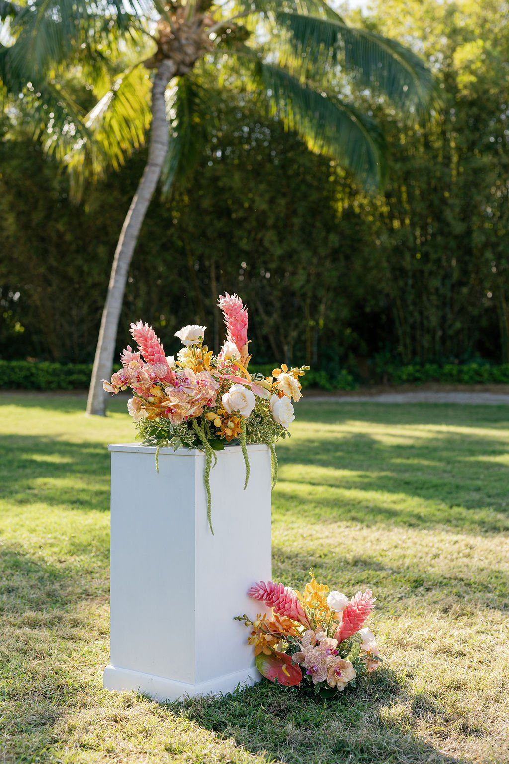 Colorful flower arrangement on a white pedestal outdoors with a palm tree and green foliage in the background.