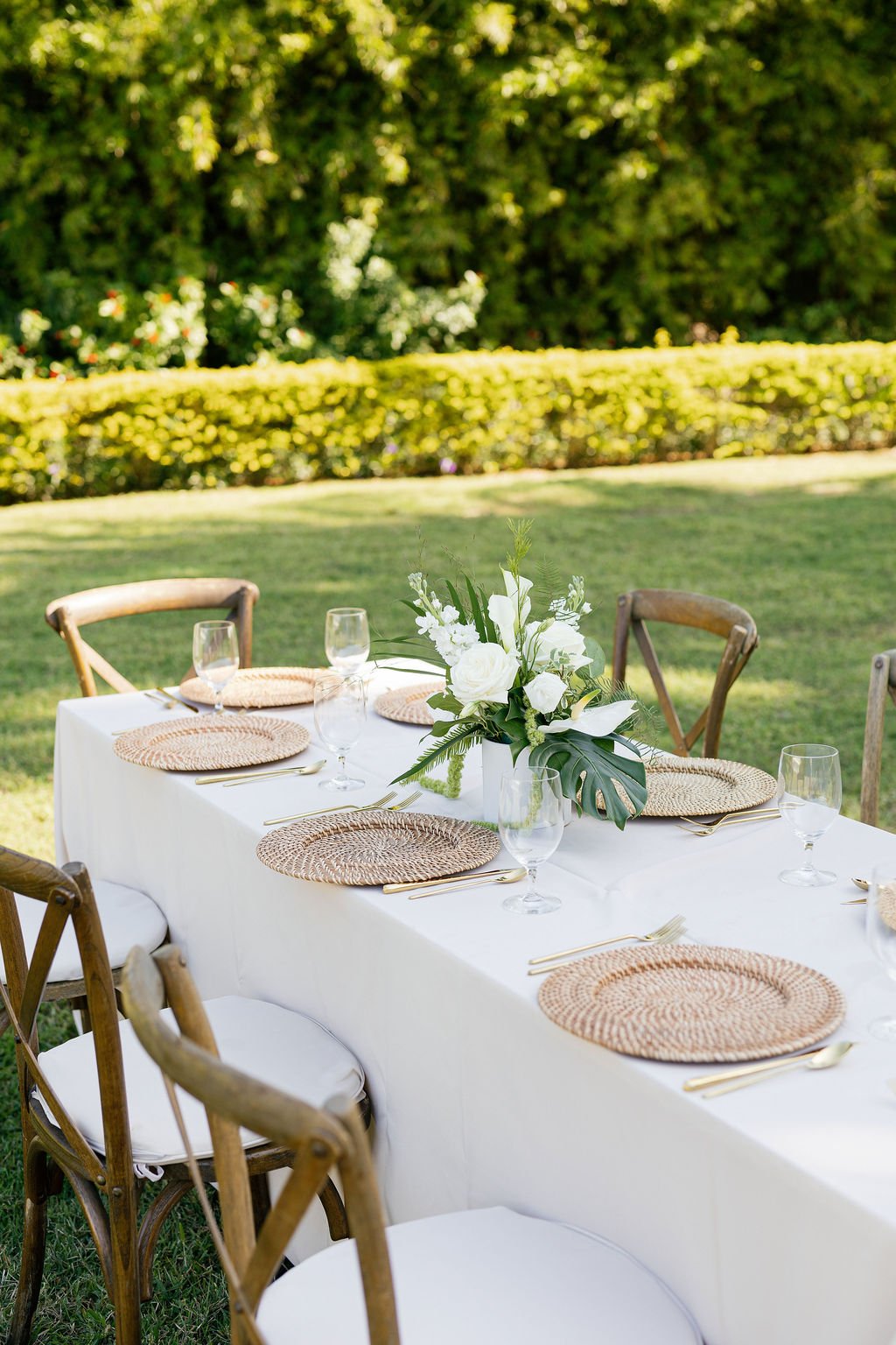 Outdoor table set for a meal with a white tablecloth, woven placemats, clear wine glasses, and a floral centerpiece with white flowers and green leaves, surrounded by wooden chairs on a grassy lawn with a lush green hedge background.