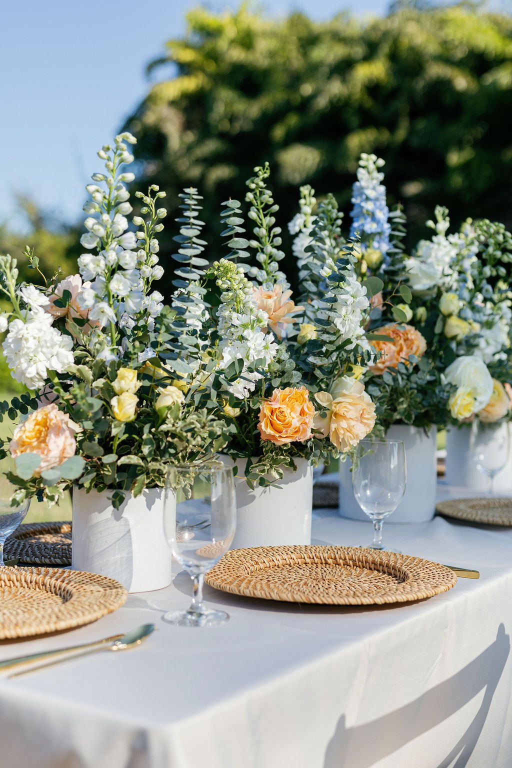 Beautiful outdoor table setup with floral centerpieces in white pots, featuring white, peach, and pastel flowers, set on a white tablecloth with woven placemats, clear wine glasses, and gold cutlery, under a sunny sky with green trees in the backgrou