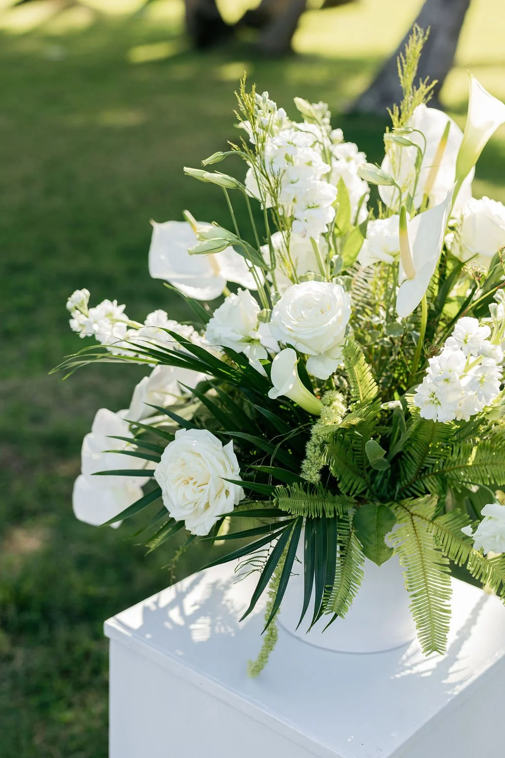 White floral arrangement with roses, lilies, and greenery on a white pedestal outdoors.