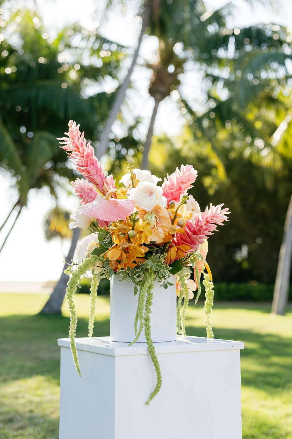 A vibrant floral arrangement with pink, white, and yellow flowers in a white vase, placed on a white pedestal outdoors with lush green trees in the background.
