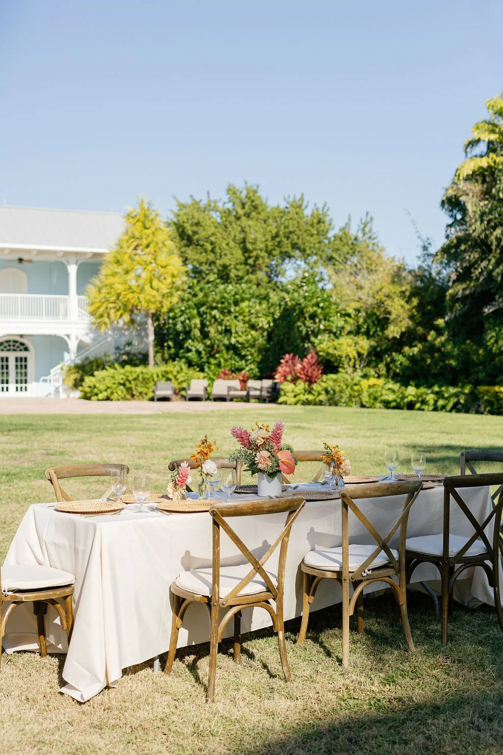 Outdoor dining table set with a white tablecloth, floral centerpiece, and glassware on a lawn, with trees, a house, and a clear blue sky in the background.