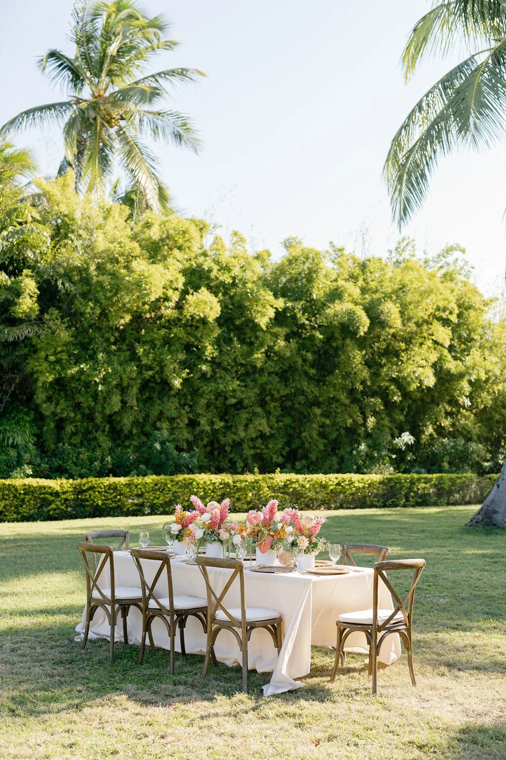 Outdoor garden setting with a long dining table decorated with pink and white floral arrangements, surrounded by wooden chairs, under tall trees and palm trees on a sunny day.