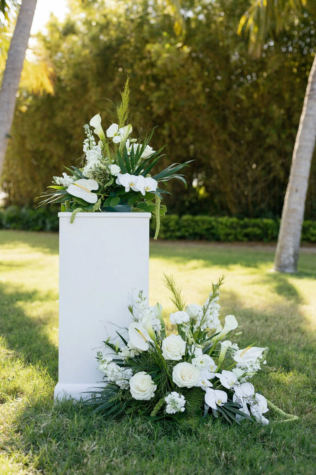 White floral arrangements with roses, calla lilies, and greenery on and beside a white pedestal outdoors with trees and grass in the background.