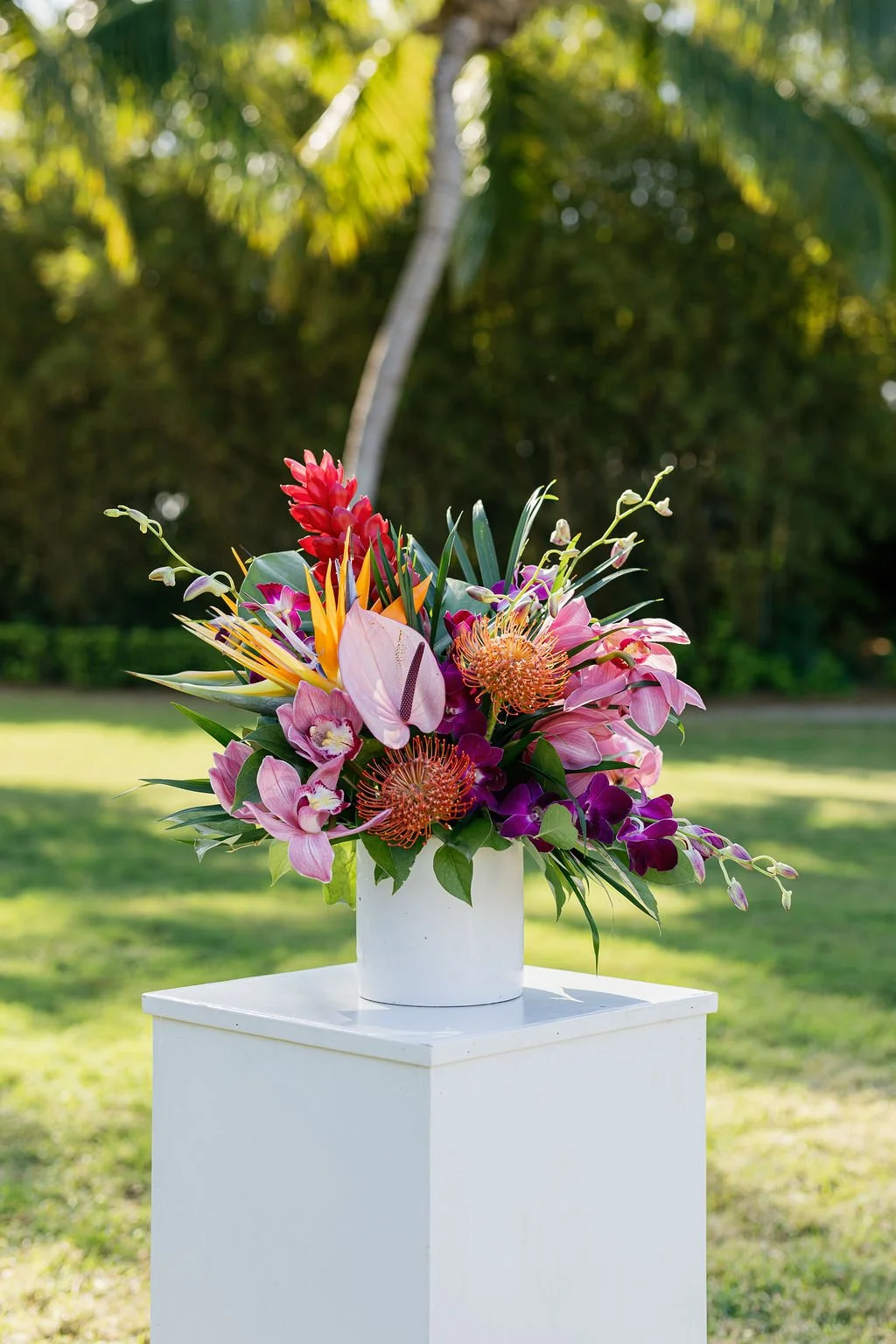 Colorful tropical flower arrangement in a white vase on a white pedestal outdoors with greenery and trees in the background.