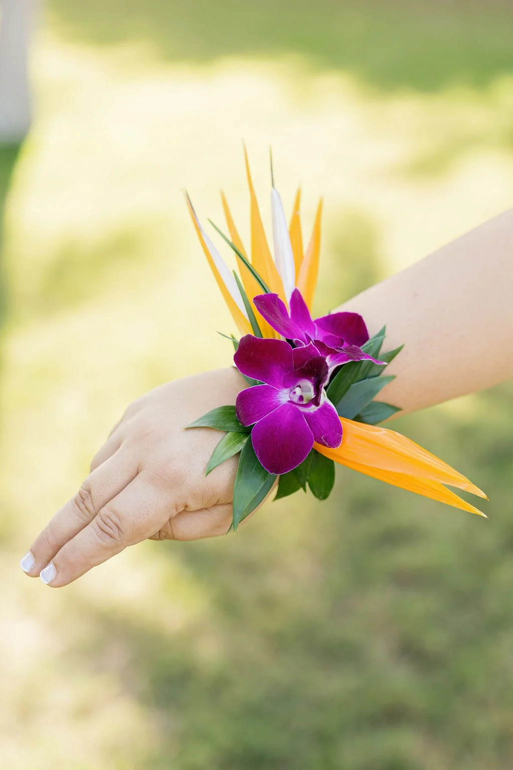 A person's wrist decorated with a colorful floral wrist corsage consisting of purple orchids, yellow bird of paradise flowers, and green leaves, with a blurred outdoor background.