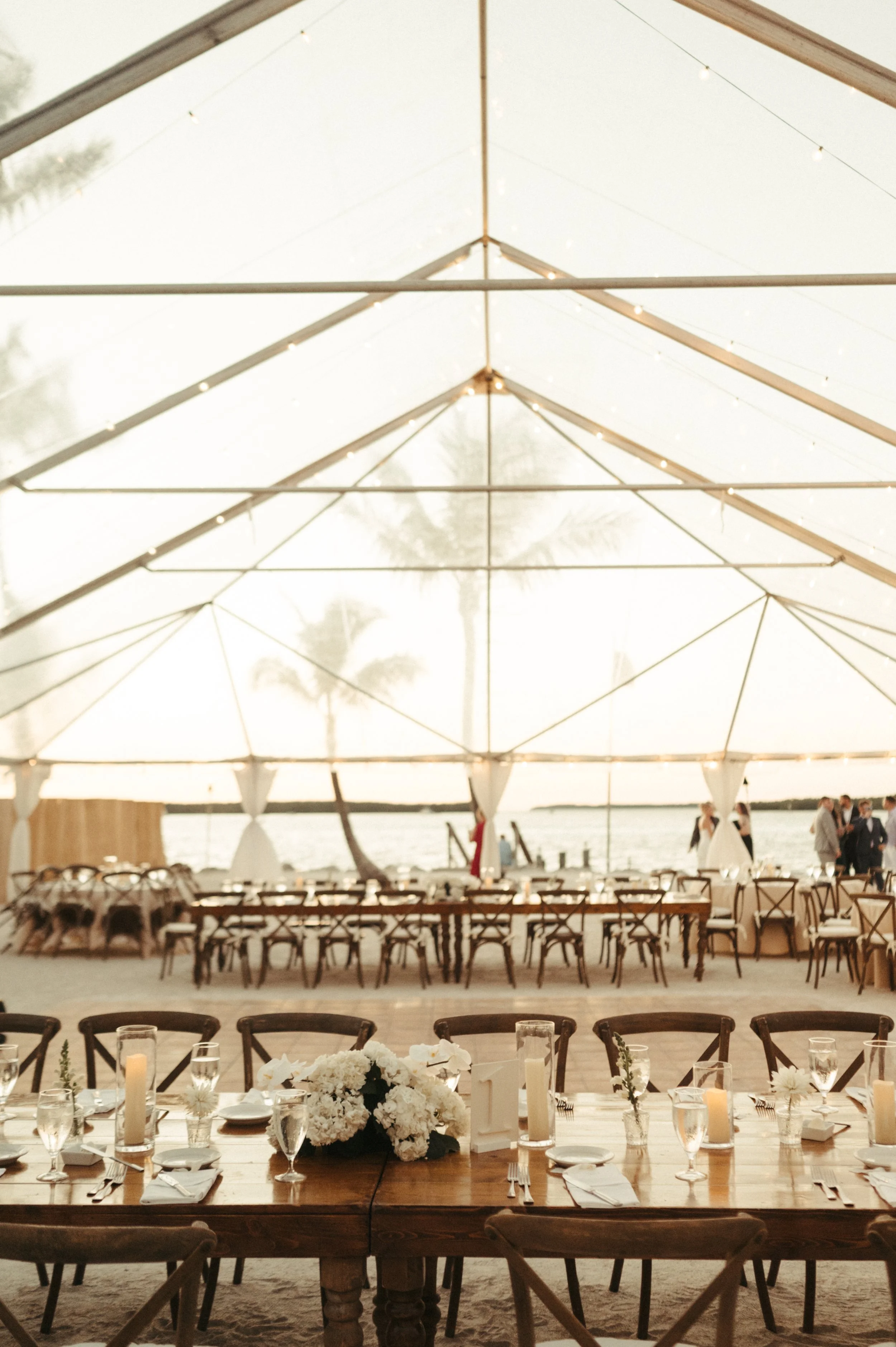 Outdoor wedding venue with wooden tables set under a clear tent, decorated with white flowers and candles, overlooking water and palm trees.